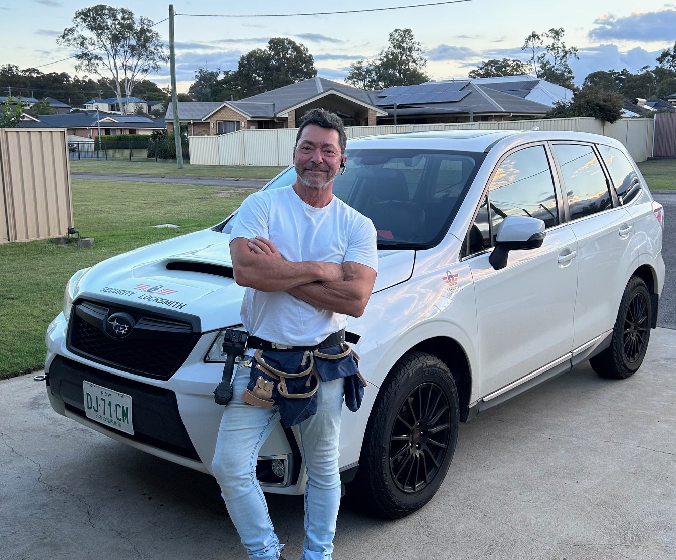 Joe standing with arms crossed leaning against the front of a white Subaru SUV with a tool belt around his waist.